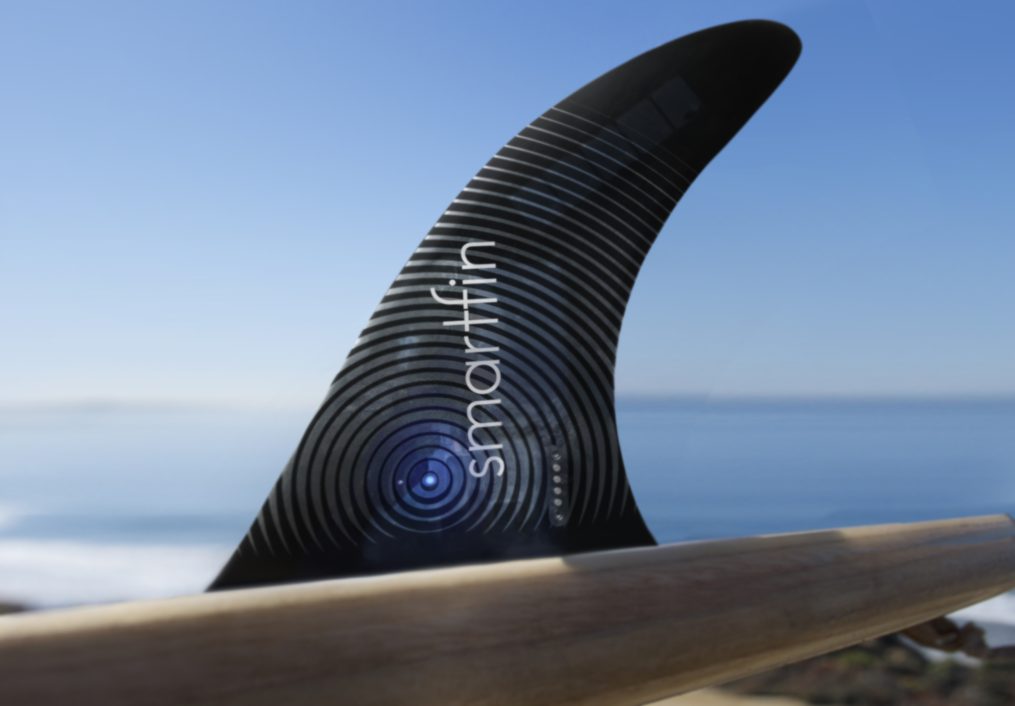 A close-up of a black surfboard fin with the Smartfin logo on it and the ocean in the background.