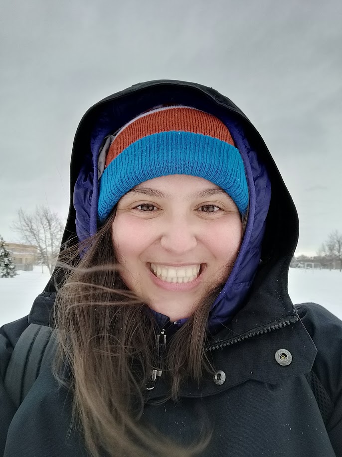 A woman with brown hair smiles at the camera. She is bundled up in a red and blue hat and a black jacket with the hood up. She's outdoors in front of a snowy background.