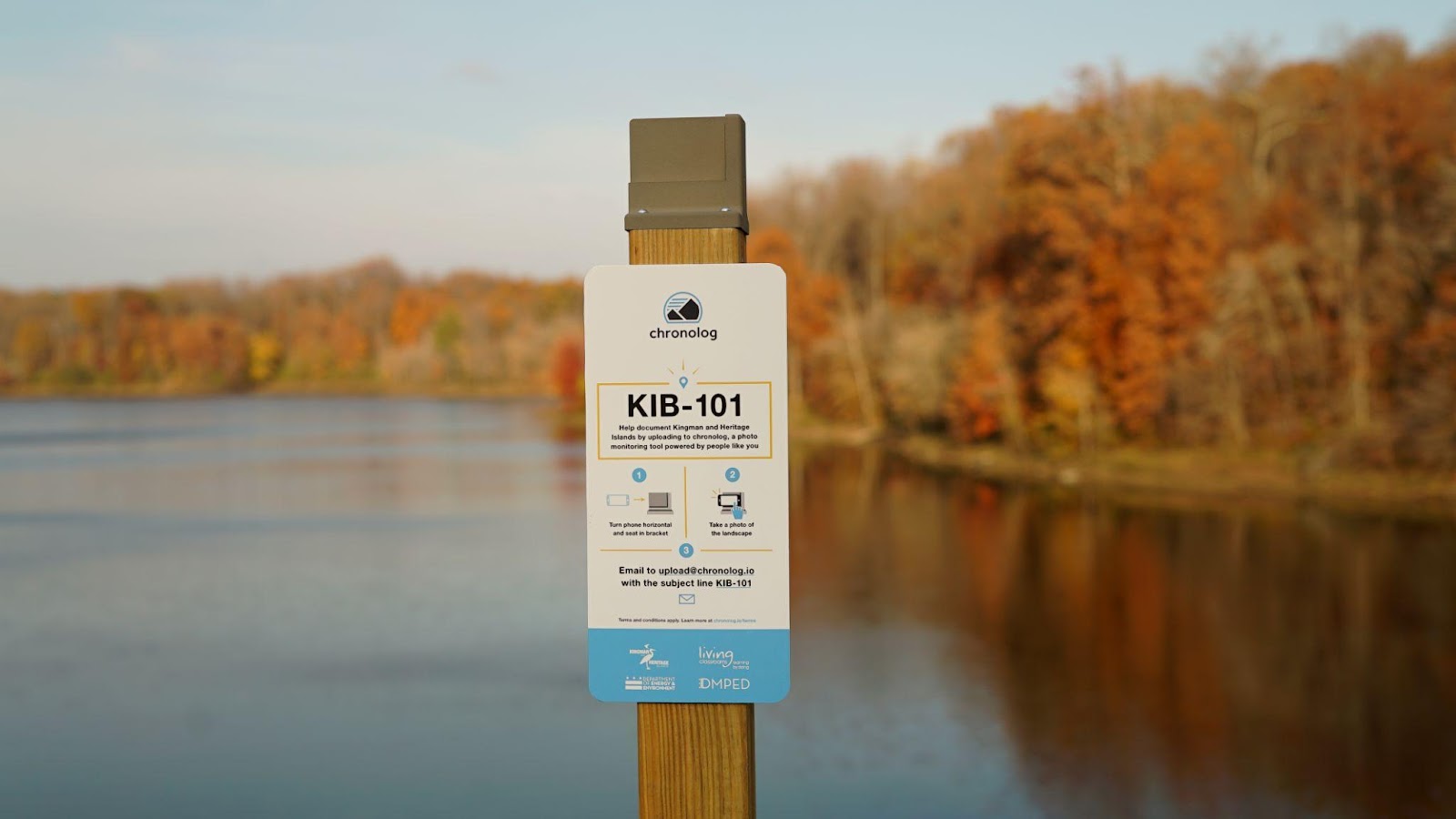 A Chronolog photo station, a post with a small sign on it, in front of a small lake surrounded by trees with fall foliage.