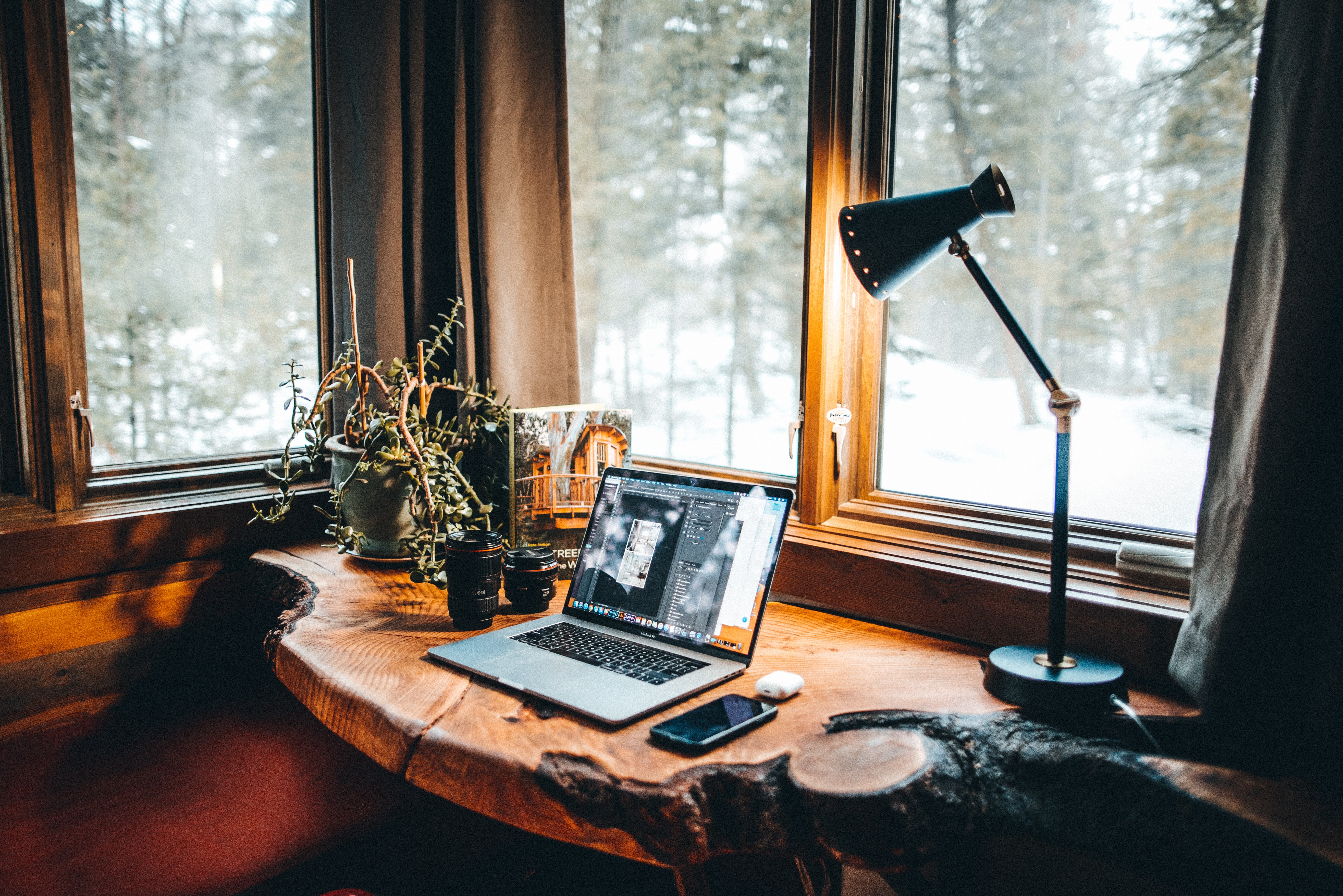 A laptop on a desk surrounded by windows overlooking the forest outside.
