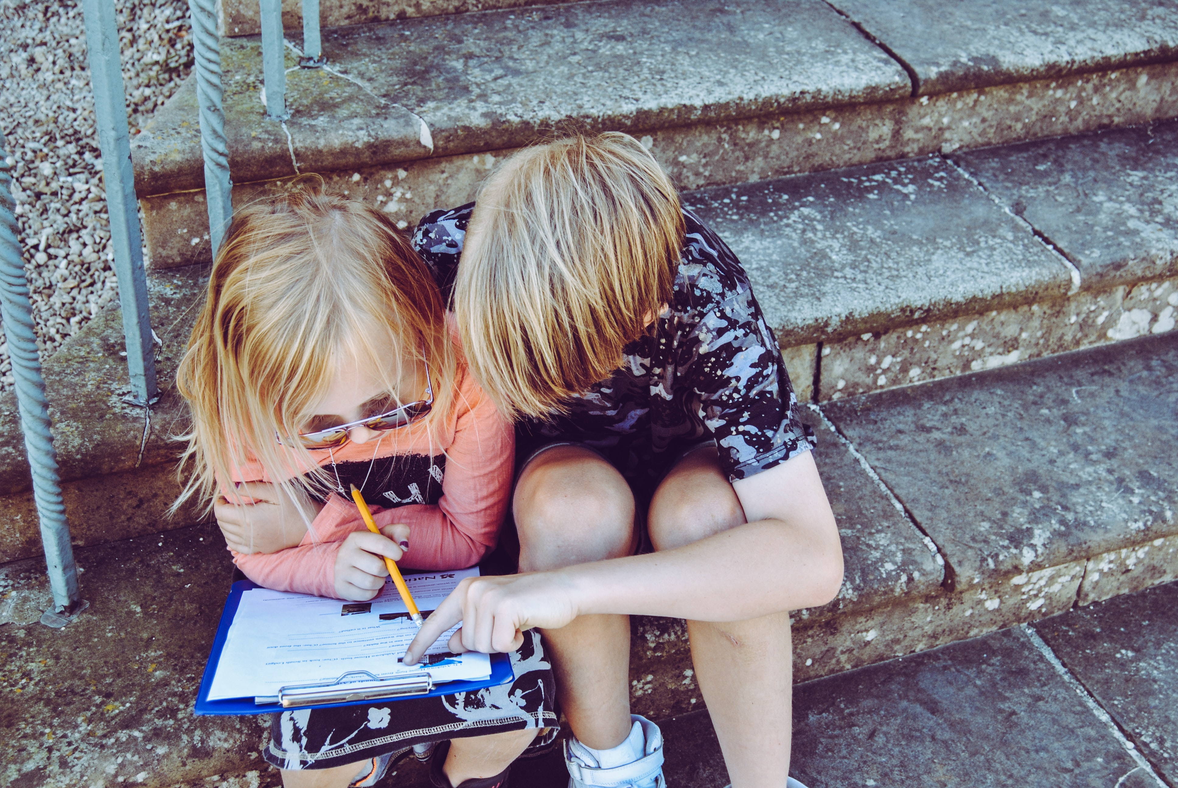 A young girl sitting on stone steps writes on a clipboard with a pencil while a young boy looks over her shoulder.