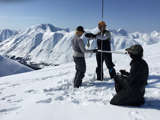 Three people in snow gear gather around a tall metal rod, a snow probe. Two are taking a measurement while the third sits with a writing utensil and paper, ready to write something down. They are standing on snow, and snow-capped mountains are visible in the near background.