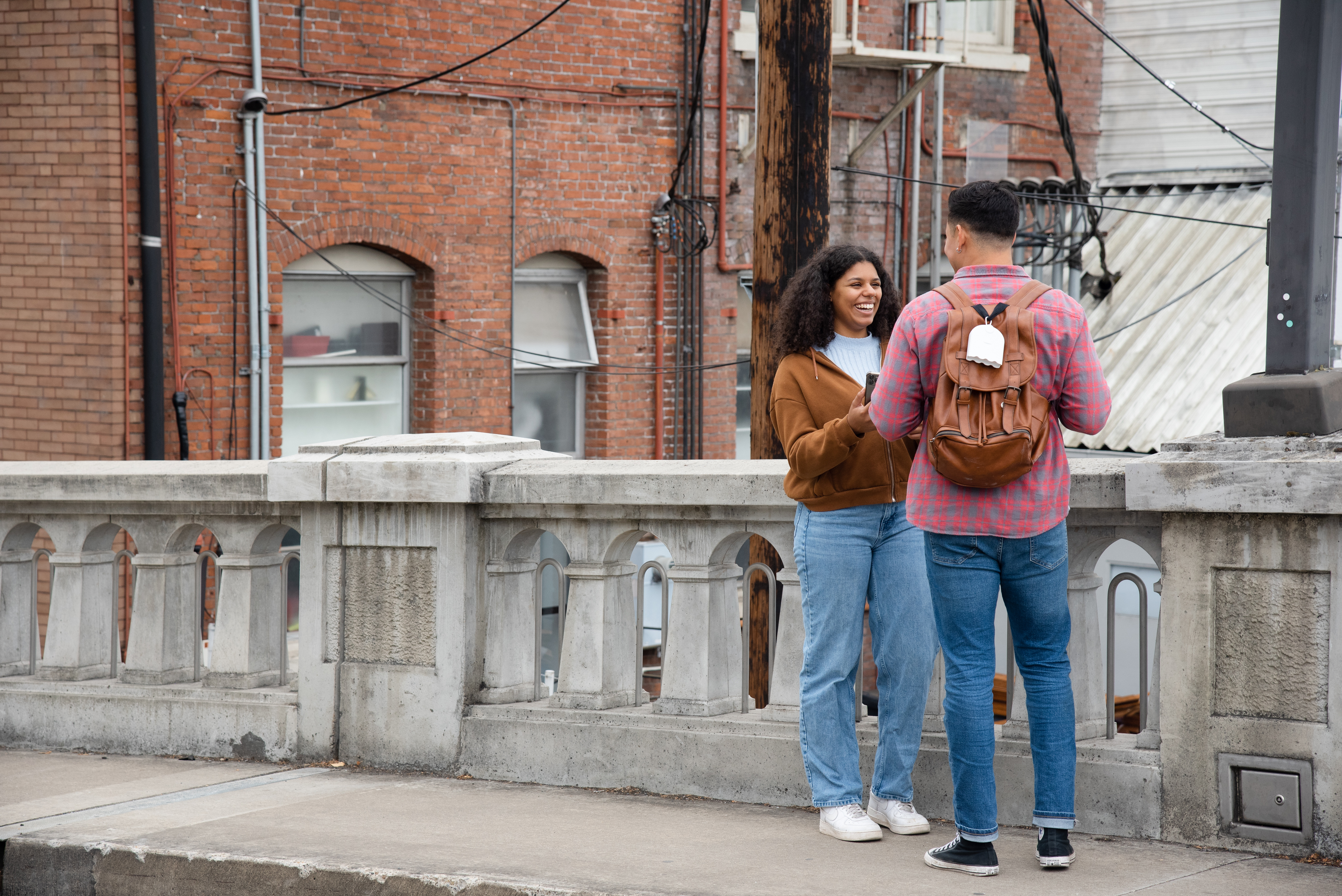 two individuals facing each other on a sidewalk. One facing away has a backpack with an air quality monitor hooked on it. The other person, facing the camera with a phone out to show use of the aircasting app.